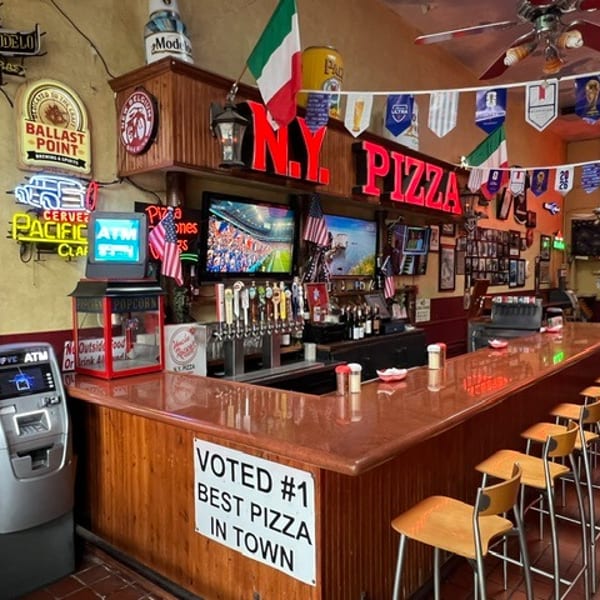 A lively pizza restaurant bar with neon signs, multiple TVs, beer taps, and bar stools.