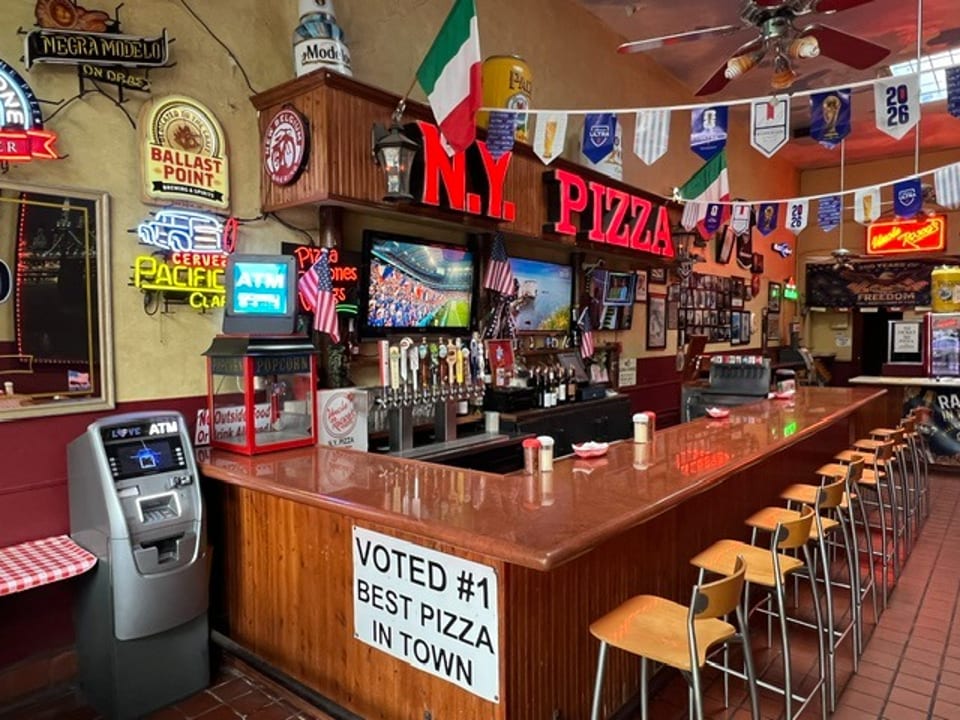 A lively pizza restaurant bar with neon signs, multiple TVs, beer taps, and bar stools.